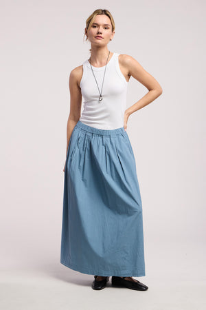 A person stands against a plain background, wearing a white sleeveless top, the CHESSA SKIRT by Velvet by Graham & Spencer—a cotton poplin A-line maxi skirt with an elastic waist—paired with black shoes and a necklace.
