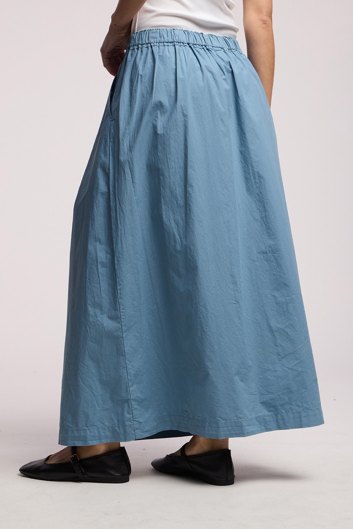 A person wears the Velvet by Graham & Spencer CHESSA SKIRT, a light blue cotton poplin maxi with an elastic waist, paired with a white top and black flats, standing against a plain background.