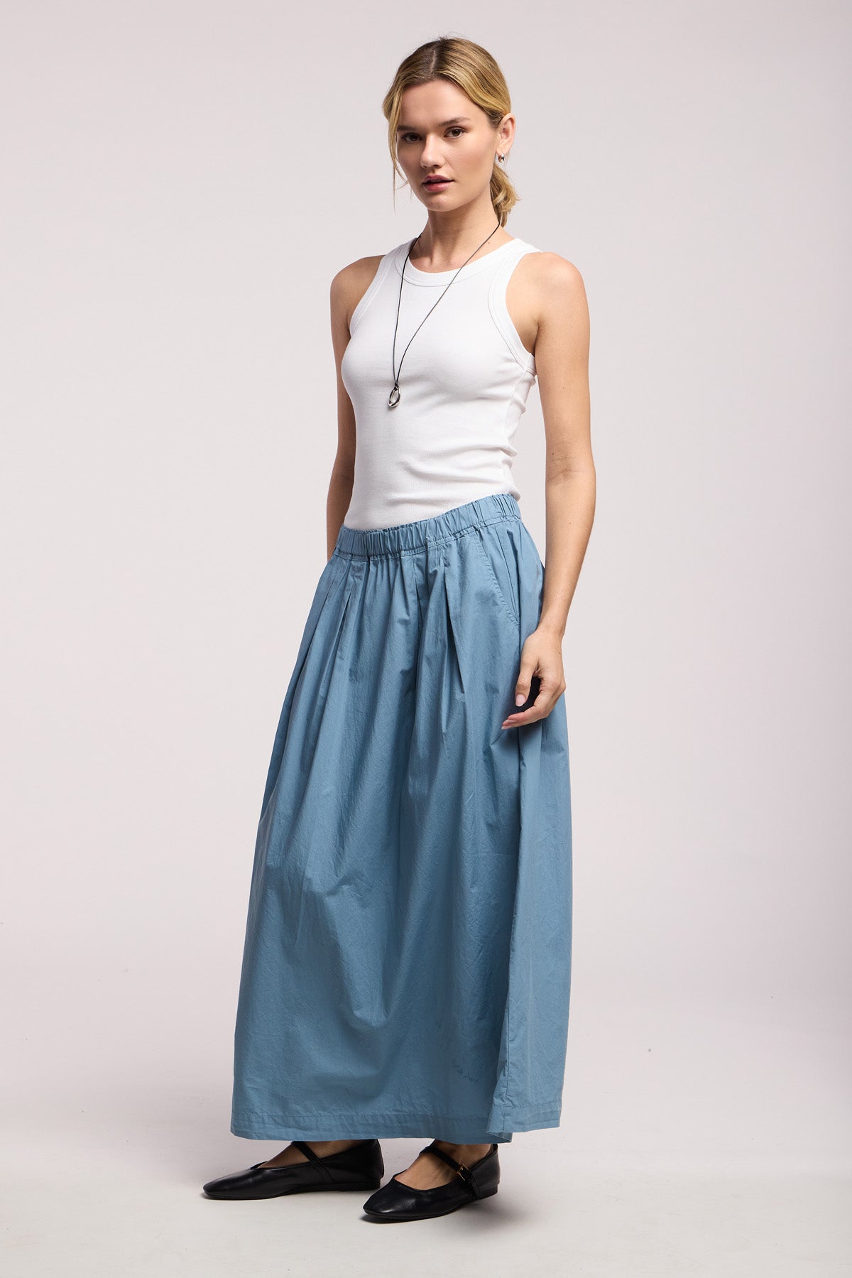 A person stands against a plain light background wearing the CHESSA SKIRT by Velvet by Graham & Spencer, a white sleeveless top, black flat shoes, and a necklace.