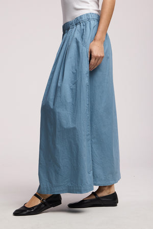 Person wearing the CHESSA SKIRT by Velvet by Graham & Spencer: a loose light blue wide-leg skirt with an elastic waist, paired with a white sleeveless top and black flat shoes, standing against a plain background.