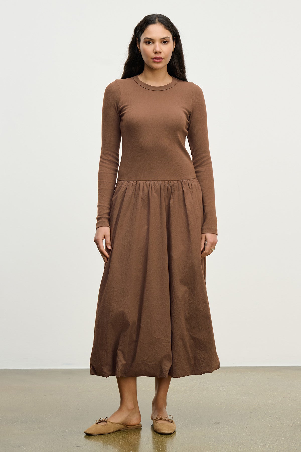 A woman stands against a plain background wearing the DOLCE DRESS by Velvet by Graham & Spencer— a long-sleeve, ribbed cotton dress with a fitted top and gathered skirt—paired with tan flat shoes.