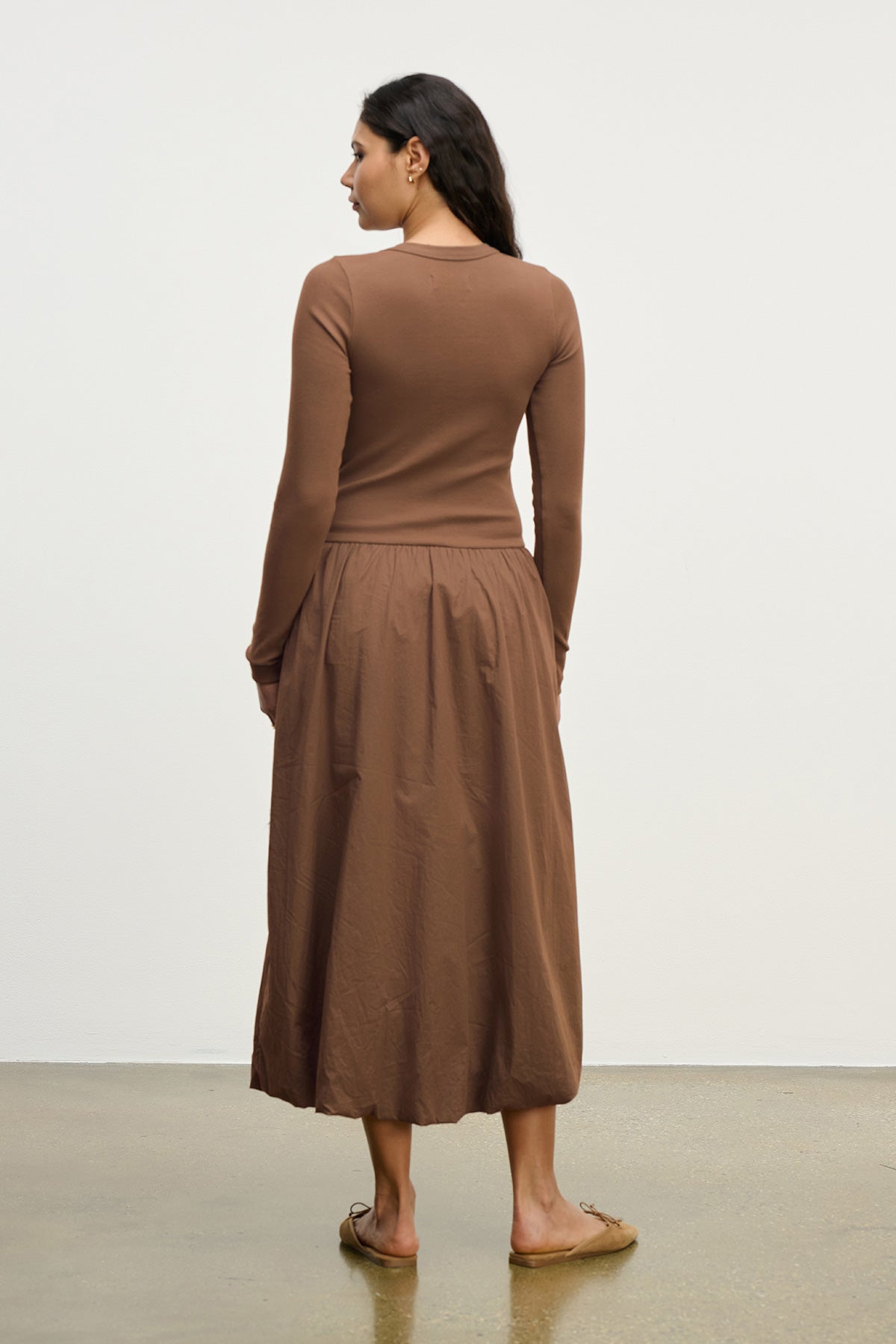 A woman stands indoors facing away, wearing the DOLCE DRESS by Velvet by Graham & Spencer. The long-sleeved brown cotton poplin dress is styled with flat shoes against a plain, light-colored background.-40248971002049