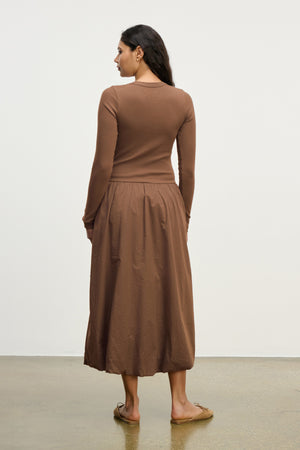 A woman stands indoors facing away, wearing the DOLCE DRESS by Velvet by Graham & Spencer. The long-sleeved brown cotton poplin dress is styled with flat shoes against a plain, light-colored background.