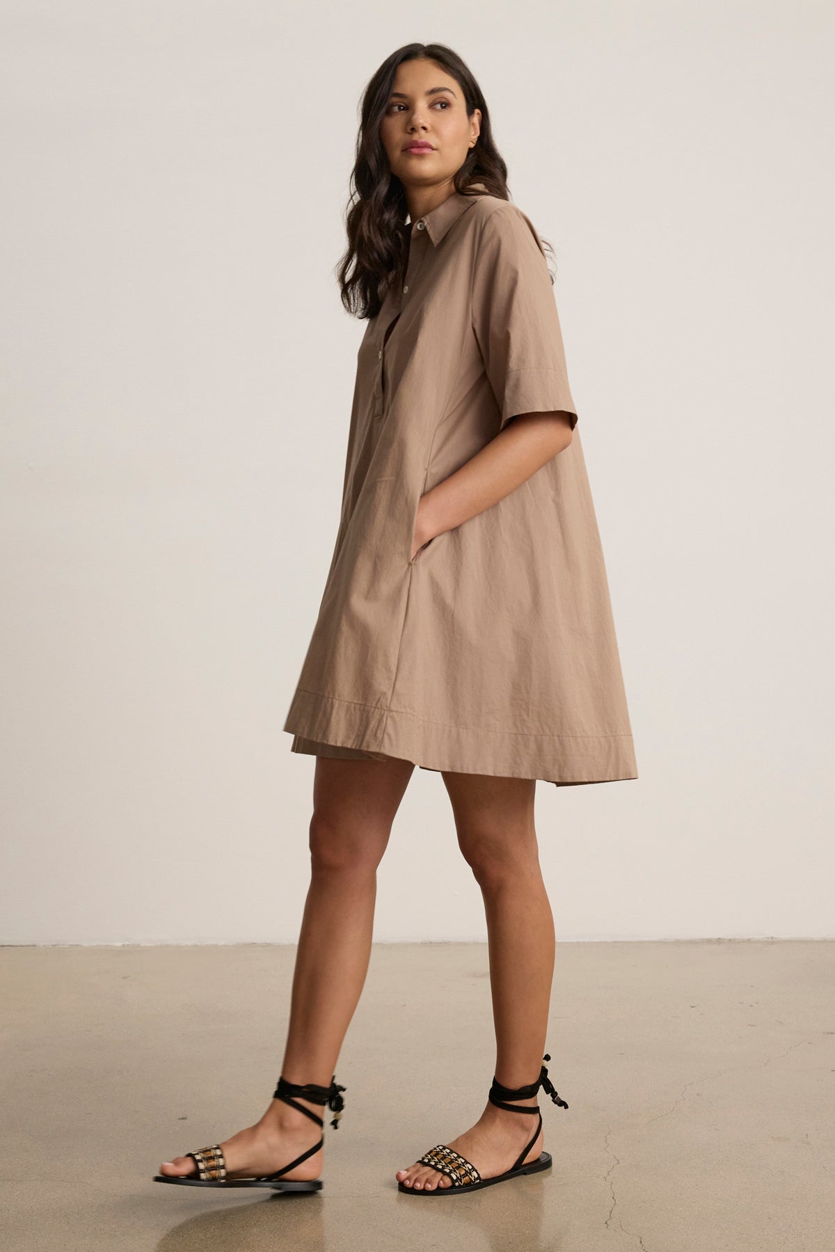 A woman wears the DRIANA DRESS by Velvet by Graham & Spencer—a loose, tan cotton poplin style with front and side seam pockets—paired with black sandals, set against a plain background.-39148705185985