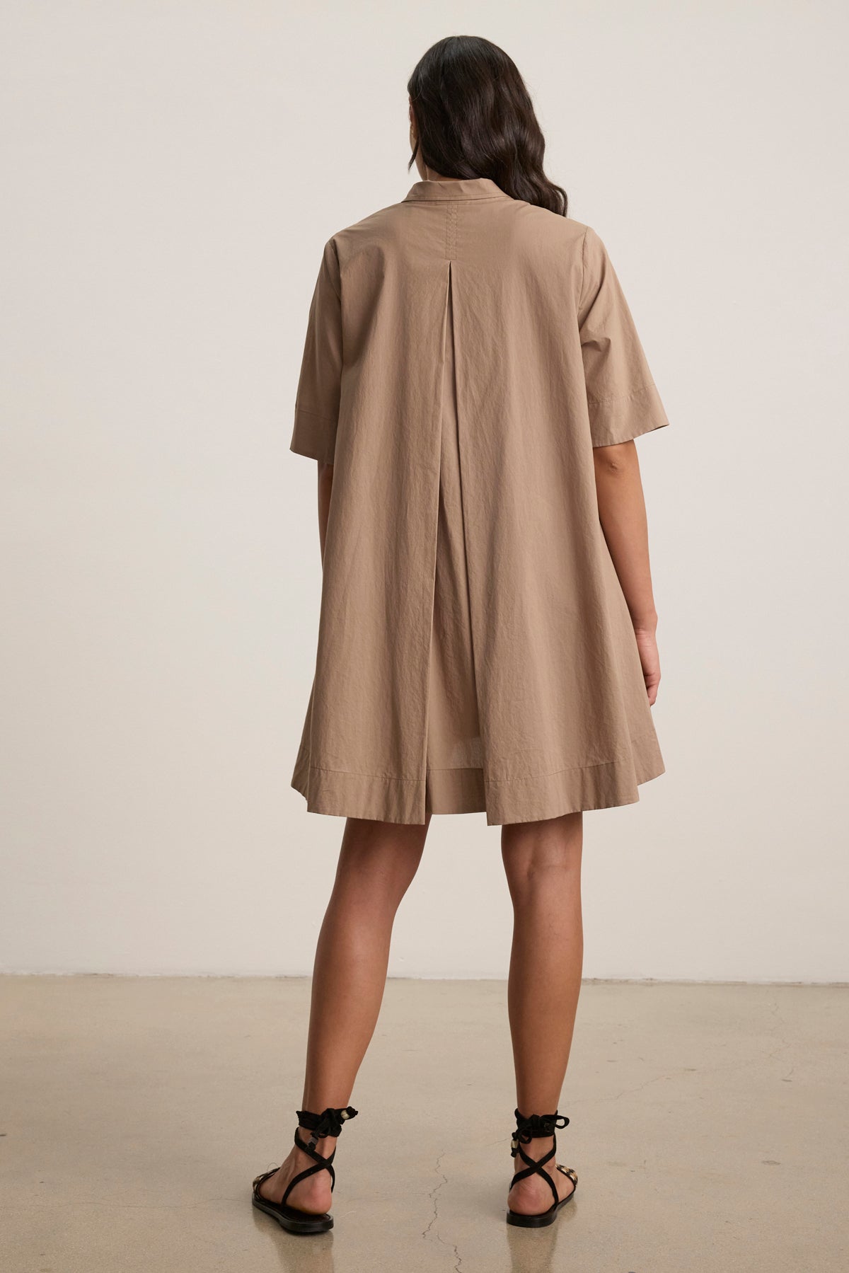 A person stands indoors facing away, wearing the DRIANA DRESS by Velvet by Graham & Spencer—a loose, knee-length, short-sleeve tan cotton poplin dress—paired with black sandals.-39148705153217
