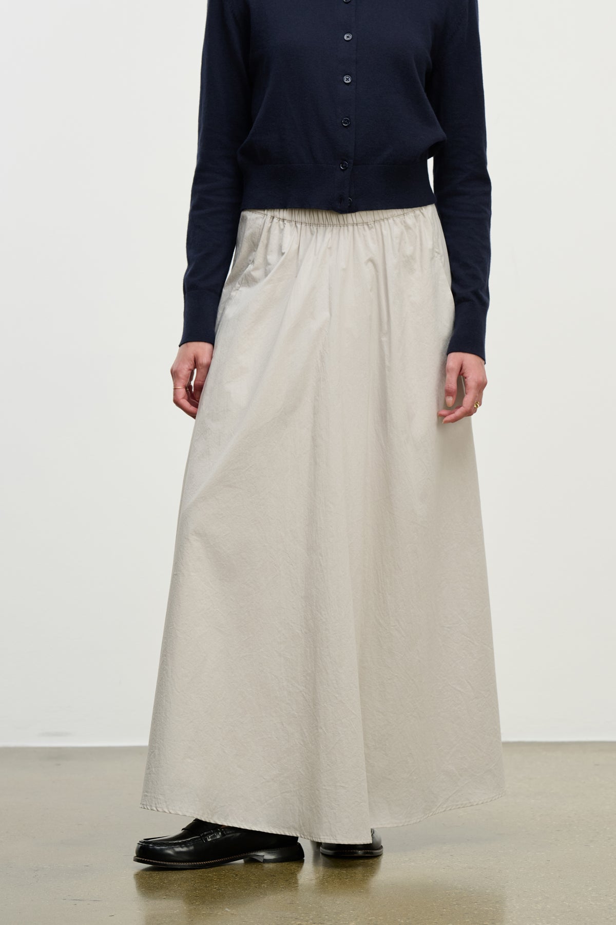 A person wears a navy long-sleeve buttoned top, the FILLA SKIRT by Velvet by Graham & Spencer in light beige cotton poplin with an elastic waistband, and black shoes, standing against a plain background.-39800308203713