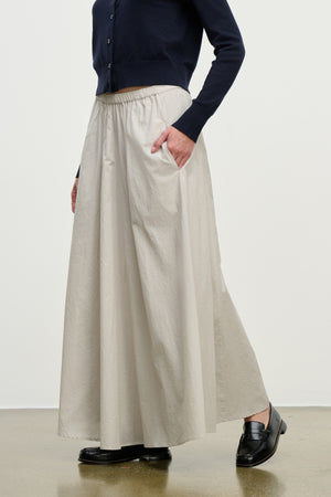 Wearing a navy long-sleeve top, the model pairs black loafers with the FILLA SKIRT by Velvet by Graham & Spencer—a beige cotton poplin skirt featuring pockets and an elastic waist—while standing against a plain background.