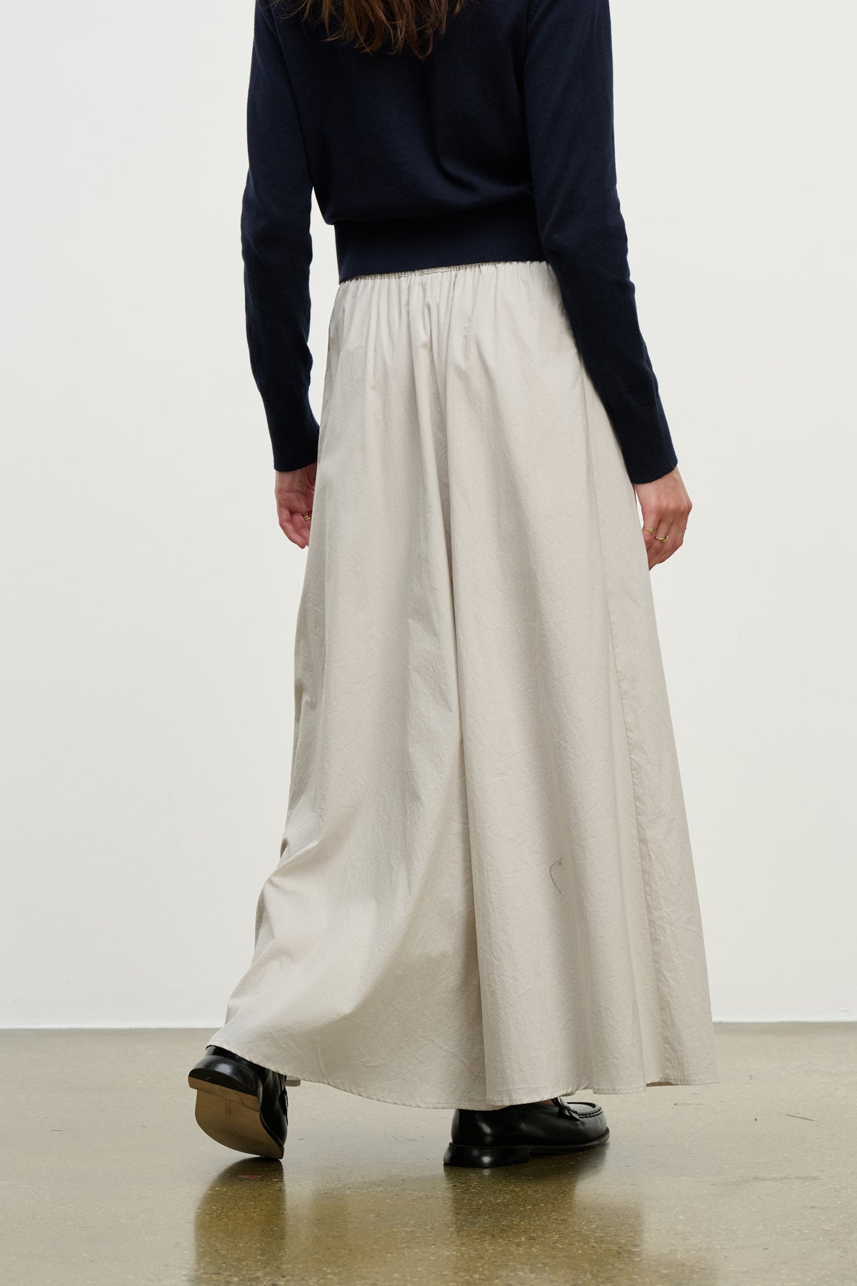 A person stands indoors in a navy long-sleeve top, black shoes, and the FILLA SKIRT—a light beige cotton poplin skirt with an elastic waist from Velvet by Graham & Spencer—on a polished floor.-39800308138177