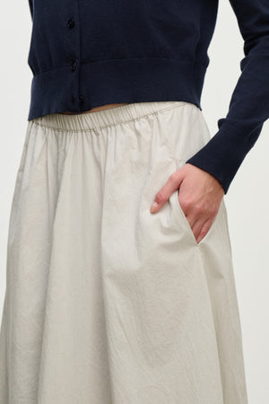 A person wearing a navy blue buttoned sweater and the FILLA SKIRT by Velvet by Graham & Spencer in light beige cotton poplin, with one hand in the skirt pocket.