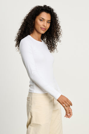 Person with curly hair wears the LOTTA TEE by Velvet by Graham & Spencer—a fitted white ribbed cotton long sleeve tee—paired with light beige pants, standing against a plain light background.