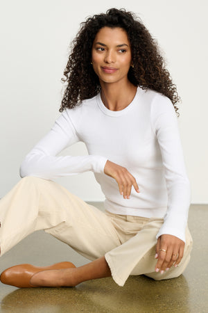 A woman with curly hair sits on the floor wearing the LOTTA TEE by Velvet by Graham & Spencer, along with beige pants and brown flats, gazing to the side with a neutral expression.