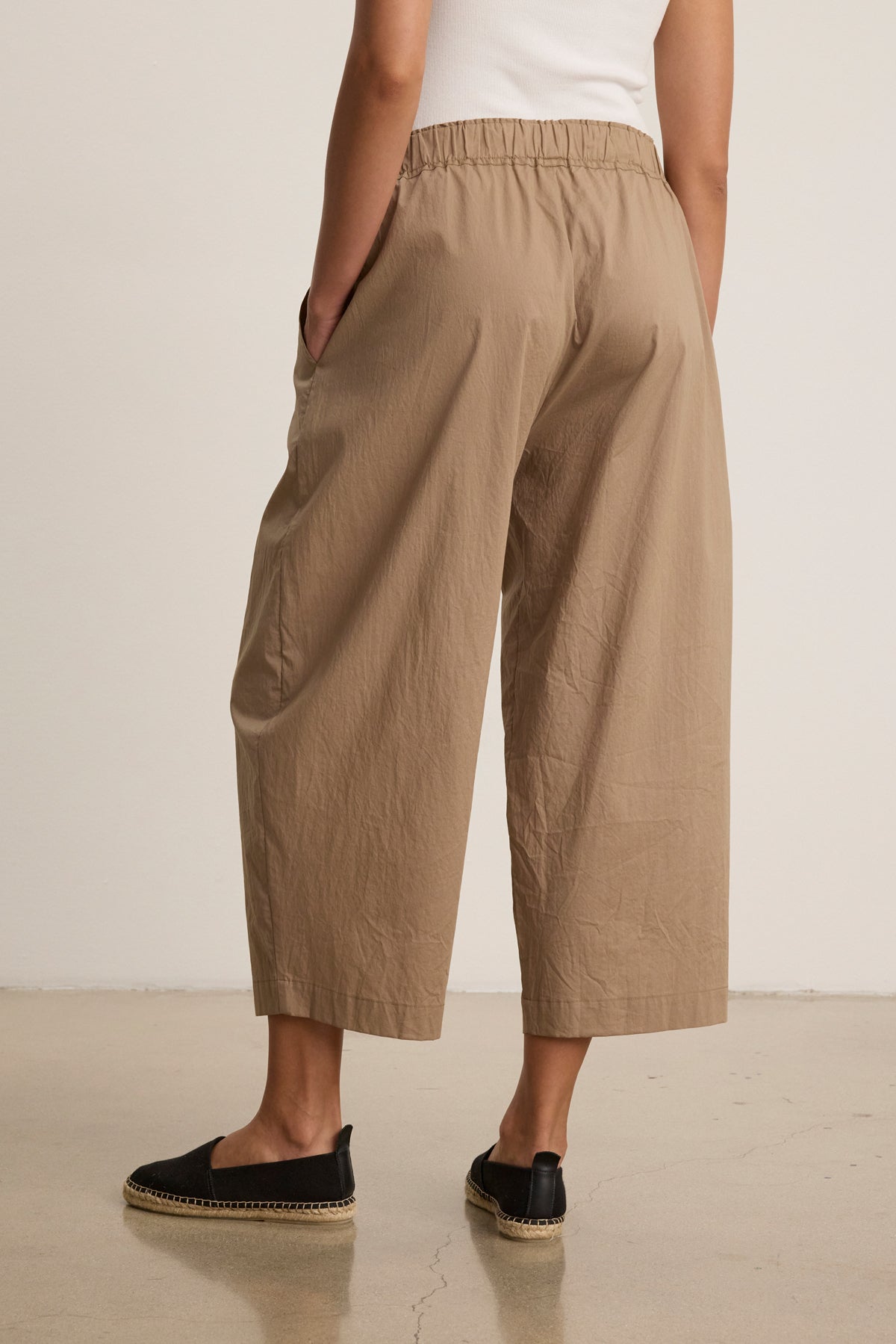 A person stands indoors, shown from the back, wearing the SOPHIA PANT in beige cotton poplin by Velvet by Graham & Spencer, styled with a white top and black espadrille shoes.-40030496129217