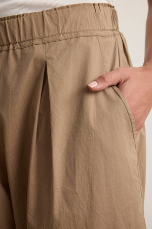 Close-up of a person wearing the SOPHIA PANT by Velvet by Graham & Spencer—beige, pleated cropped balloon pants with an elastic waistband and side pockets. A soft white cotton poplin shirt peeks out, creating a relaxed silhouette.