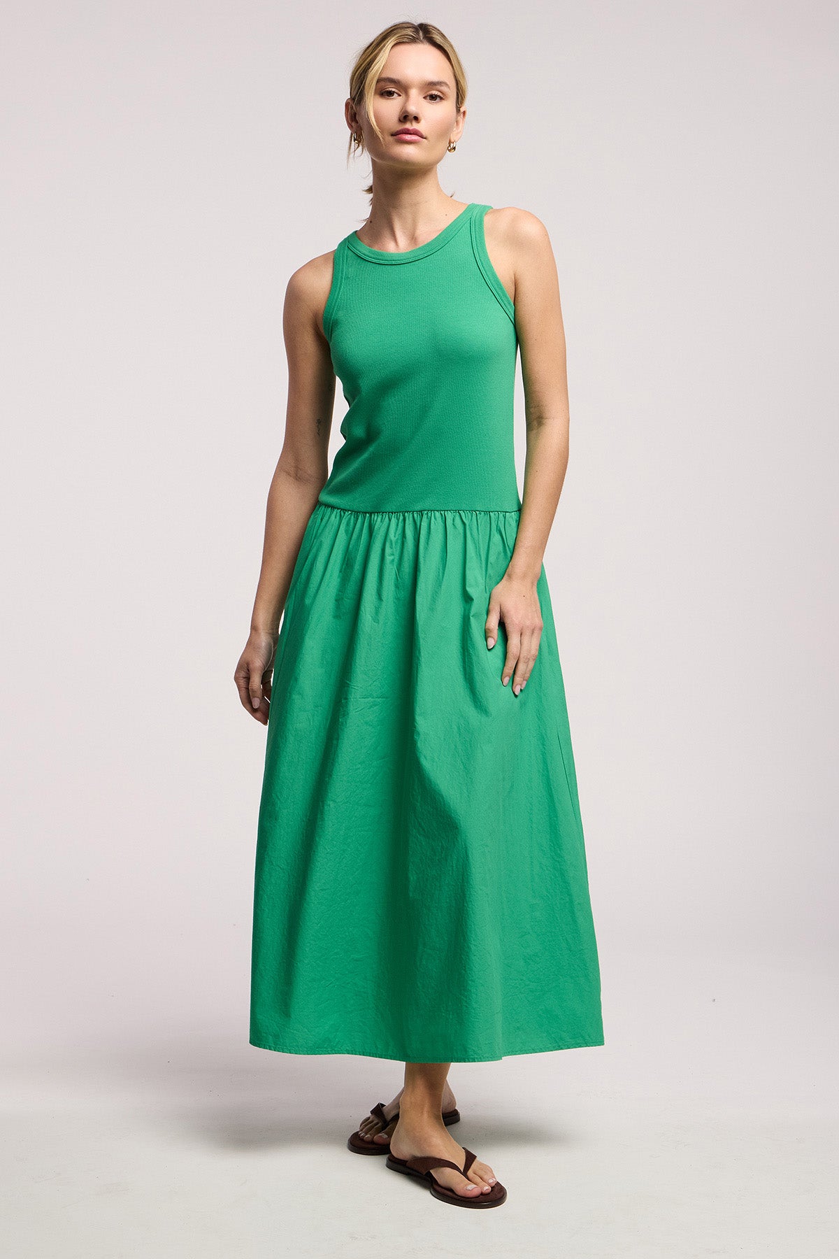 A woman wears the ROMILLY DRESS by Velvet by Graham & Spencer—a sleeveless green cotton poplin dress with a fitted ribbed bodice—and brown flip-flops, standing against a plain light background.-41527503749313
