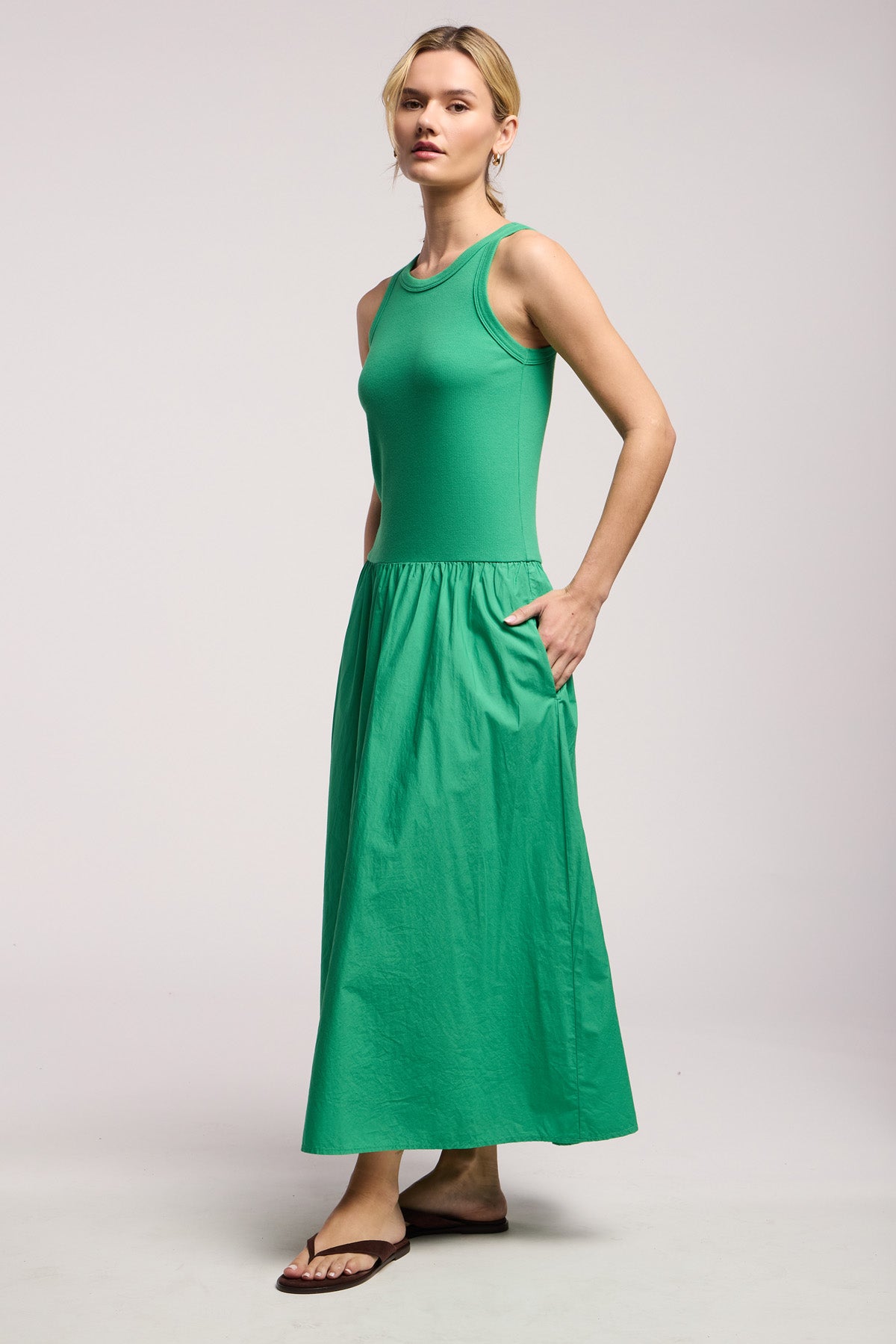 A woman wears the Velvet by Graham & Spencer ROMILLY DRESS—a sleeveless green cotton poplin maxi with pockets—and black sandals, set against a plain light background.