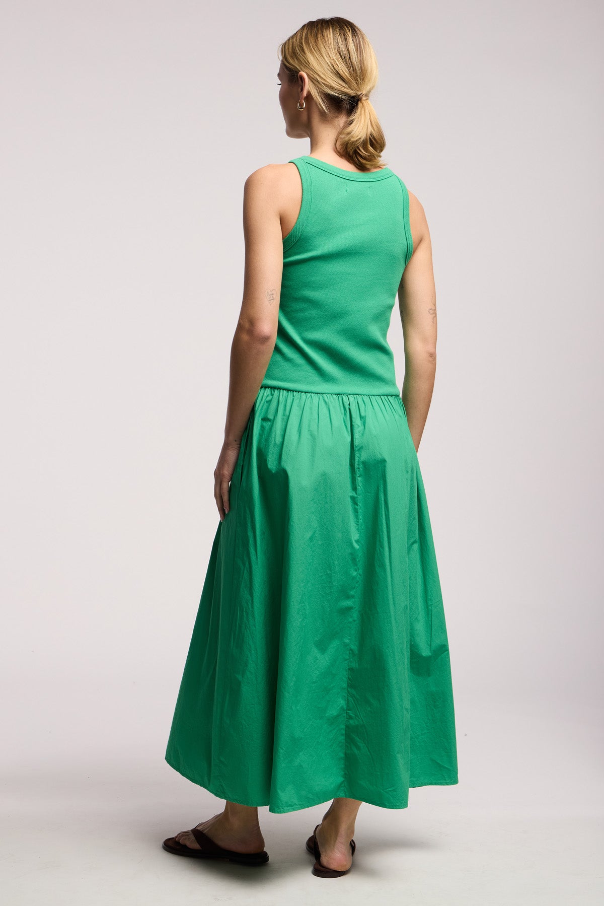A person stands facing away, wearing the ROMILLY DRESS by Velvet by Graham & Spencer—a green drop waist dress with a fitted ribbed bodice and matching cotton poplin skirt—paired with black sandals, against a plain light background.