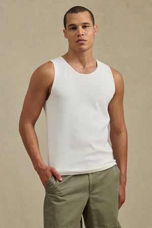 A person stands against a plain background wearing the CARLOS TANK by Velvet by Graham & Spencer, a white ribbed cotton crewneck tank, paired with olive green shorts and one hand in a pocket.