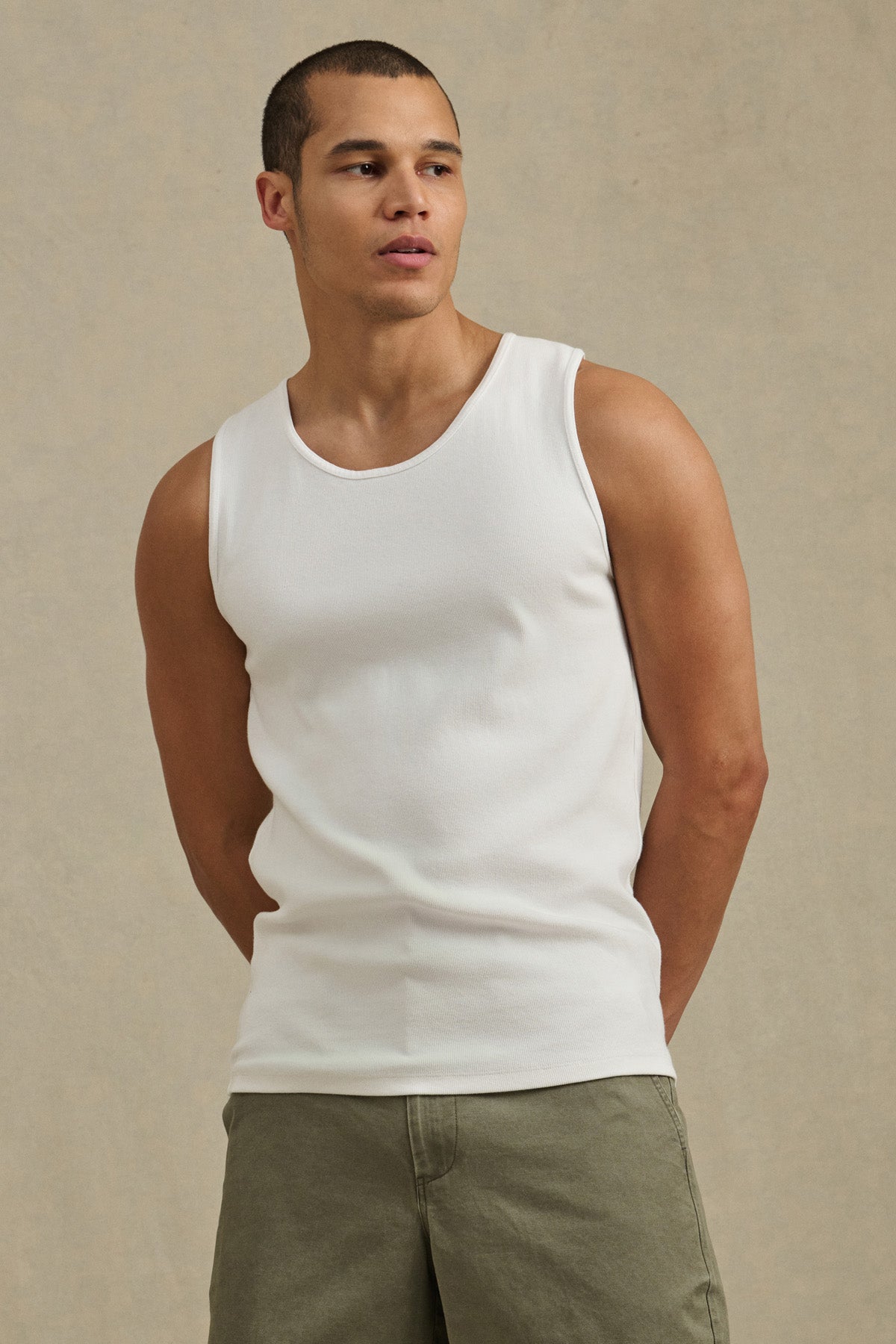 A man in the CARLOS TANK by Velvet by Graham & Spencer, featuring a classic crew neckline and ribbed cotton design, pairs it with olive green shorts against a plain beige background.-41428090421441
