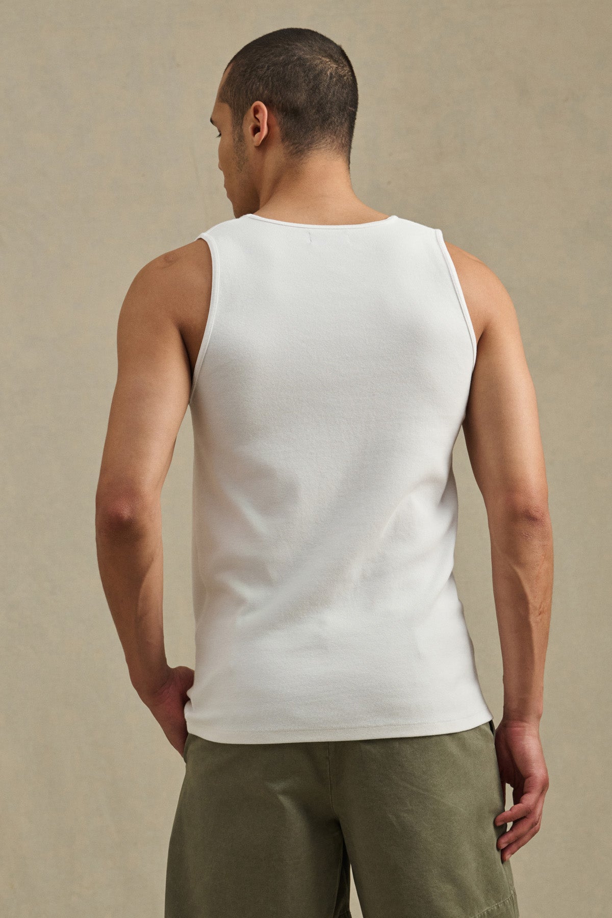 A man with short hair stands with his back to the camera, wearing the Velvet by Graham & Spencer CARLOS TANK in white and olive green shorts against a plain background.