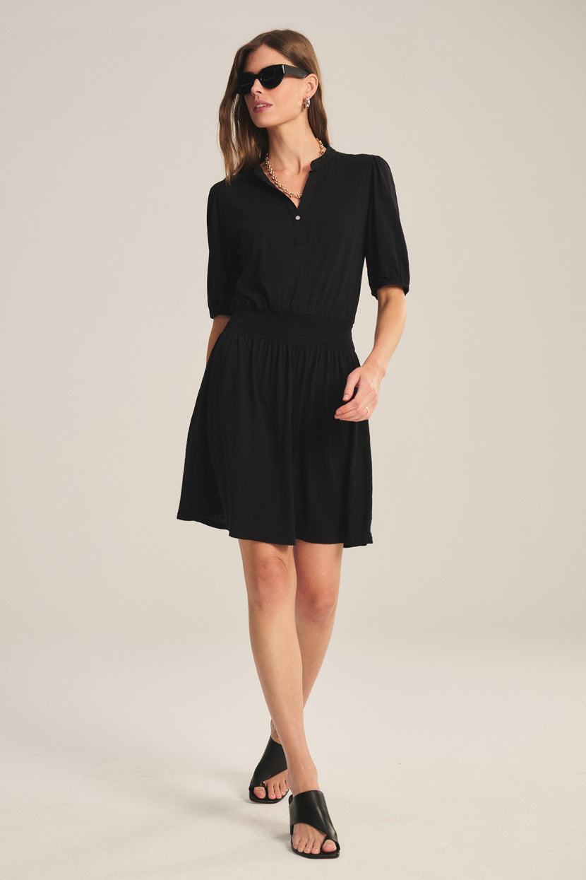 A woman wearing the RIDLEY DRESS by Velvet by Graham & Spencer— a V-neck black mini made from lightweight cotton slub—black sunglasses, and black slide sandals stands against a plain light background.