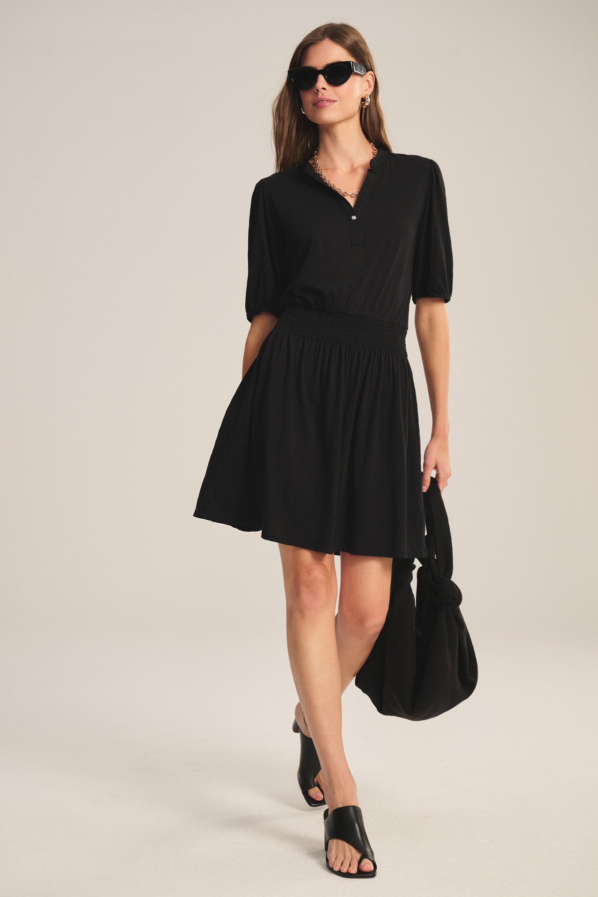 A woman stands against a light background wearing the RIDLEY DRESS by Velvet by Graham & Spencer—a black V-neck cotton slub mini dress—paired with black sandals, sunglasses, and a black bag.-41338484129985
