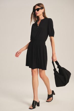 Woman in the RIDLEY DRESS by Velvet by Graham & Spencer, paired with black heeled sandals, sunglasses, and a large black bag, walks against a plain background.