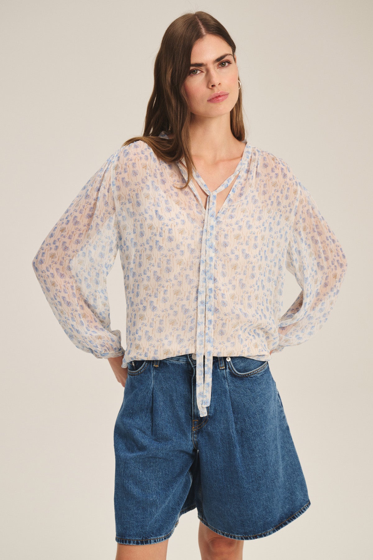 A woman poses against a plain background wearing the RIELA TOP by Velvet by Graham & Spencer, a sheer long-sleeve cami with a floral print, paired with blue denim shorts.