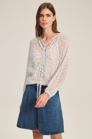 A woman wears the Velvet by Graham & Spencer RIELA TOP, a light printed viscose blouse with a tie neckline, paired with high-waisted denim shorts, standing against a plain background.