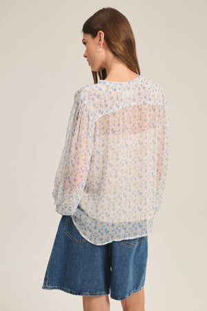A woman with long brown hair wears the RIELA TOP by Velvet by Graham & Spencer, a sheer cami in a light floral print, paired with blue denim shorts and facing away from the camera against a plain background.