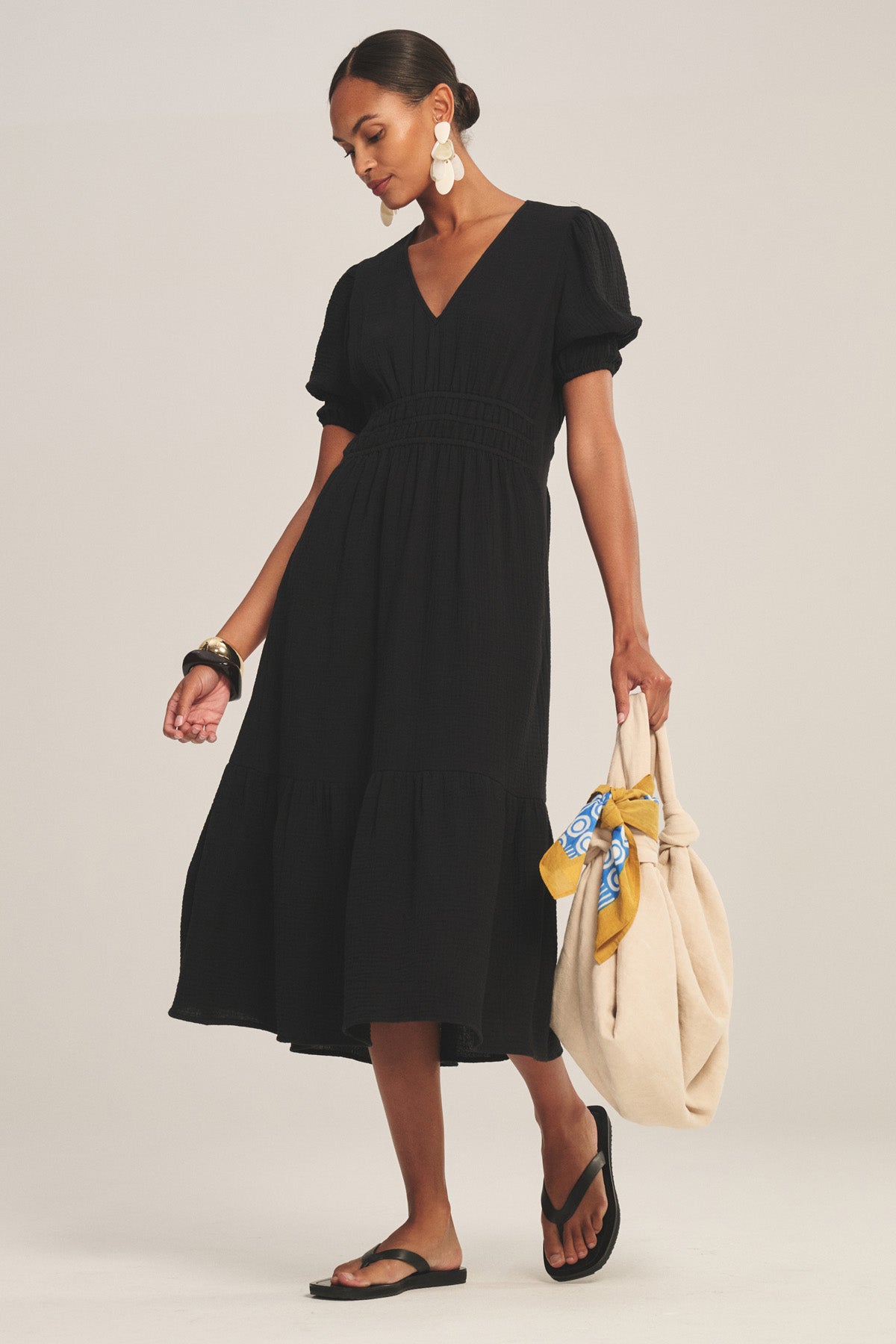 A woman wears the RIVELLE DRESS by Velvet by Graham & Spencer, a black cotton gauze midi with short puff sleeves and v-neck. She holds a beige bag with a yellow scarf, paired with black sandals and large white earrings.