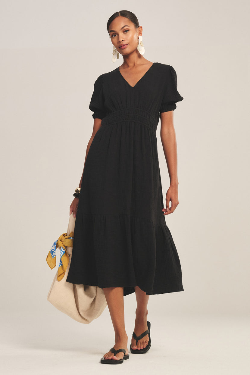 A woman stands wearing the RIVELLE DRESS by Velvet by Graham & Spencer— a black cotton gauze midi dress with puff sleeves—styled with black sandals, large white earrings, and a beige tote bag adorned with a yellow patterned scarf.
