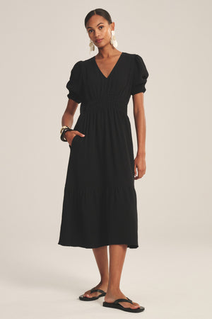 A woman wears the RIVELLE DRESS by Velvet by Graham & Spencer—a black, short-sleeved cotton gauze midi with a V-neck, puff sleeves, and tiered skirt—paired with black sandals, a bracelet, and large earrings.