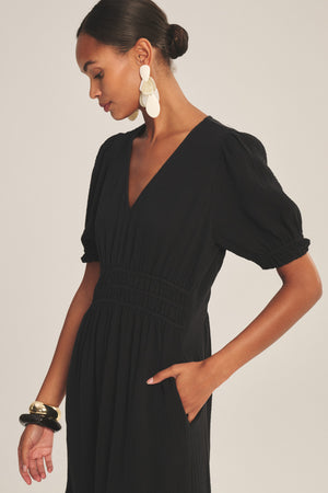 A woman models the RIVELLE DRESS by Velvet by Graham & Spencer—a black puff sleeve, V-neck design—paired with statement earrings and chunky bracelets, posing with one hand in her pocket against a plain background.