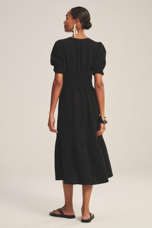A woman faces away, wearing the RIVELLE DRESS by Velvet by Graham & Spencer—a black cotton gauze midi dress with a gathered waist and skirt—styled with a bun, earrings, bracelets, and black sandals for an effortlessly chic look.