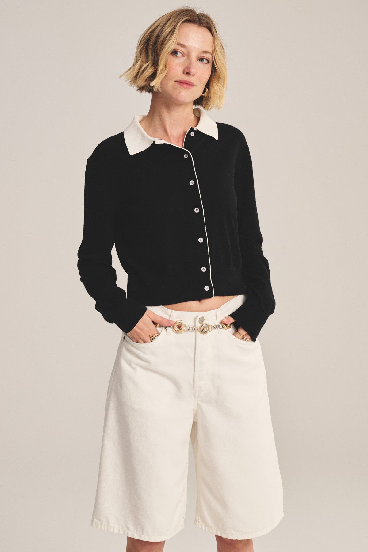 A woman stands with hands in pockets, wearing the RIVEN CARDIGAN by Velvet by Graham & Spencer—a black cotton-cashmere button-up with a white collar—paired with white knee-length shorts, against a plain background.-41044121125057