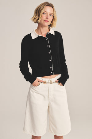 A woman stands with hands in pockets, wearing the RIVEN CARDIGAN by Velvet by Graham & Spencer—a black cotton-cashmere button-up with a white collar—paired with white knee-length shorts, against a plain background.