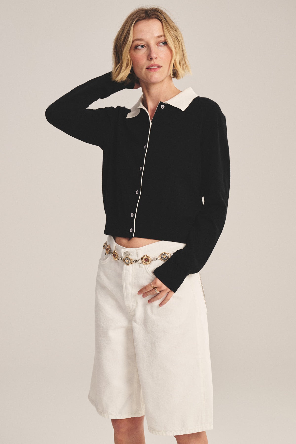 A woman wears the RIVEN CARDIGAN by Velvet by Graham & Spencer—a black cotton-cashmere cropped sweater with a white collar—paired with white knee-length shorts and a chain belt, posing against a plain background.-41044121059521
