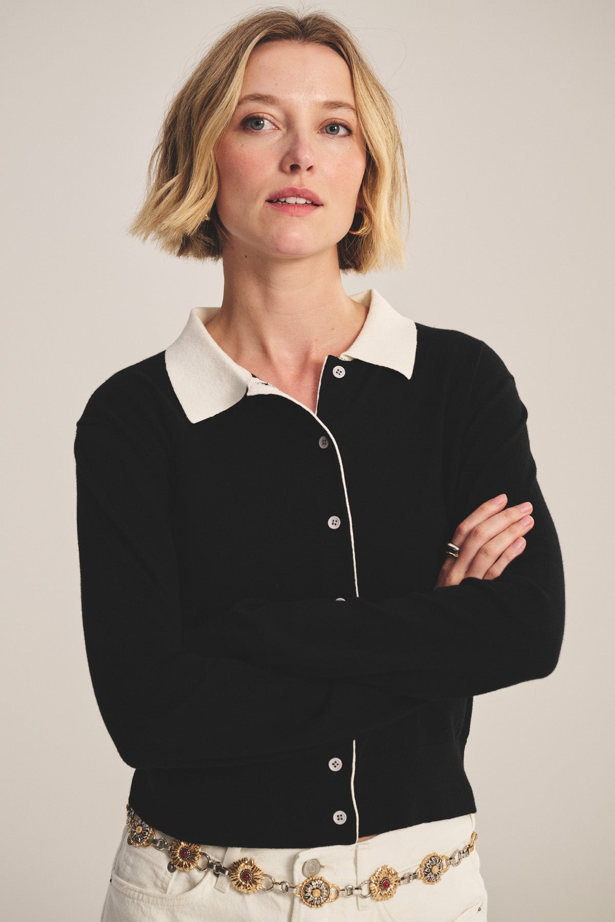A woman with short blonde hair stands with her arms crossed, wearing the RIVEN CARDIGAN by Velvet by Graham & Spencer—a cotton-cashmere sweater polo with a white collar—paired with white pants and a decorative belt.-41044120993985