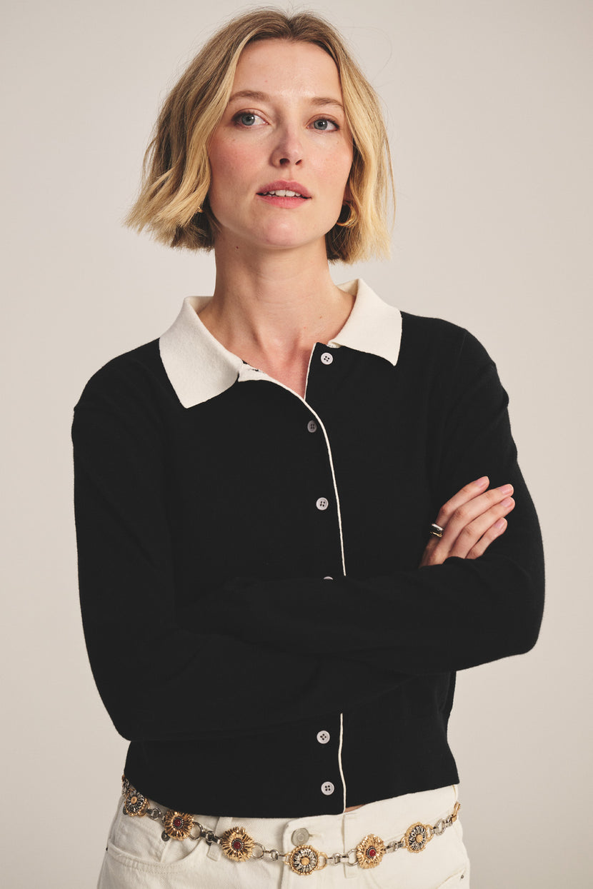 A woman with short blonde hair stands with her arms crossed, wearing the RIVEN CARDIGAN by Velvet by Graham & Spencer—a cotton-cashmere sweater polo with a white collar—paired with white pants and a decorative belt.