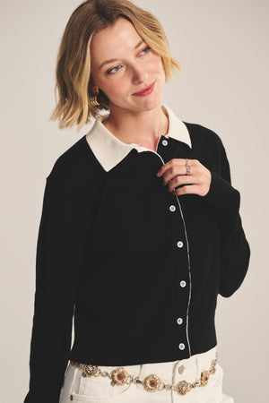A person with short blonde hair wears the RIVEN CARDIGAN by Velvet by Graham & Spencer, featuring a white collar, styled with white pants, a decorative belt, and multiple rings, standing against a neutral background.