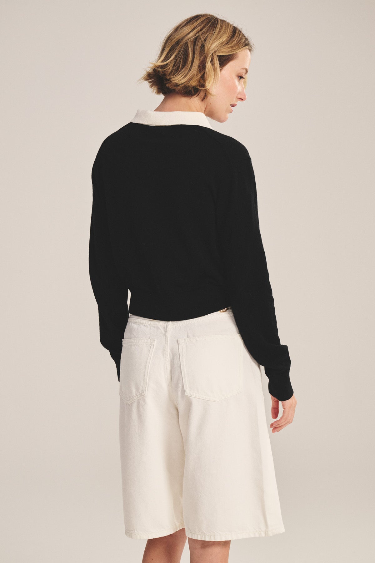 A person faces away wearing the Velvet by Graham & Spencer RIVEN CARDIGAN, a black cotton-cashmere sweater polo with a white collar, paired with white knee-length shorts.-41044120895681