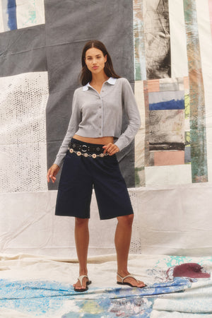 A woman stands before an abstract fabric backdrop, wearing the RIVEN CARDIGAN by Velvet by Graham & Spencer in light gray, paired with navy knee-length shorts, a decorative belt, and sandals.