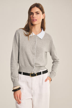 A woman wears the RIVEN CARDIGAN by Velvet by Graham & Spencer—a light gray cotton-cashmere cropped cardigan with a white collar—paired with white pants and a black belt, standing against a plain background.