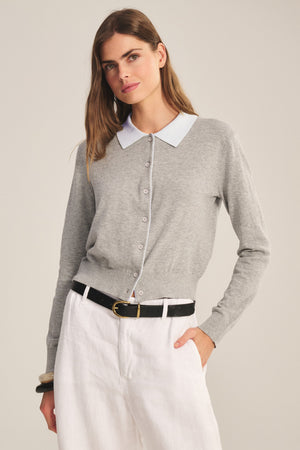 A person wears the RIVEN CARDIGAN by Velvet by Graham & Spencer—a light gray cotton-cashmere sweater polo with a white collar—paired with white pants and a black belt, standing against a plain background.