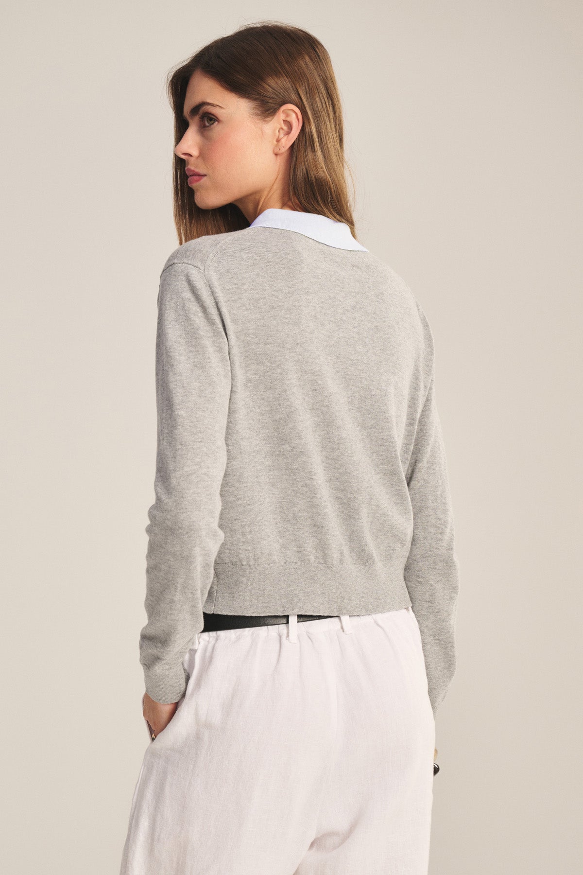 A woman with long brown hair faces away, wearing the RIVEN CARDIGAN by Velvet by Graham & Spencer—a light grey cotton-cashmere sweater polo—layered over a white collared shirt and white pants against a plain background.-41277142728897