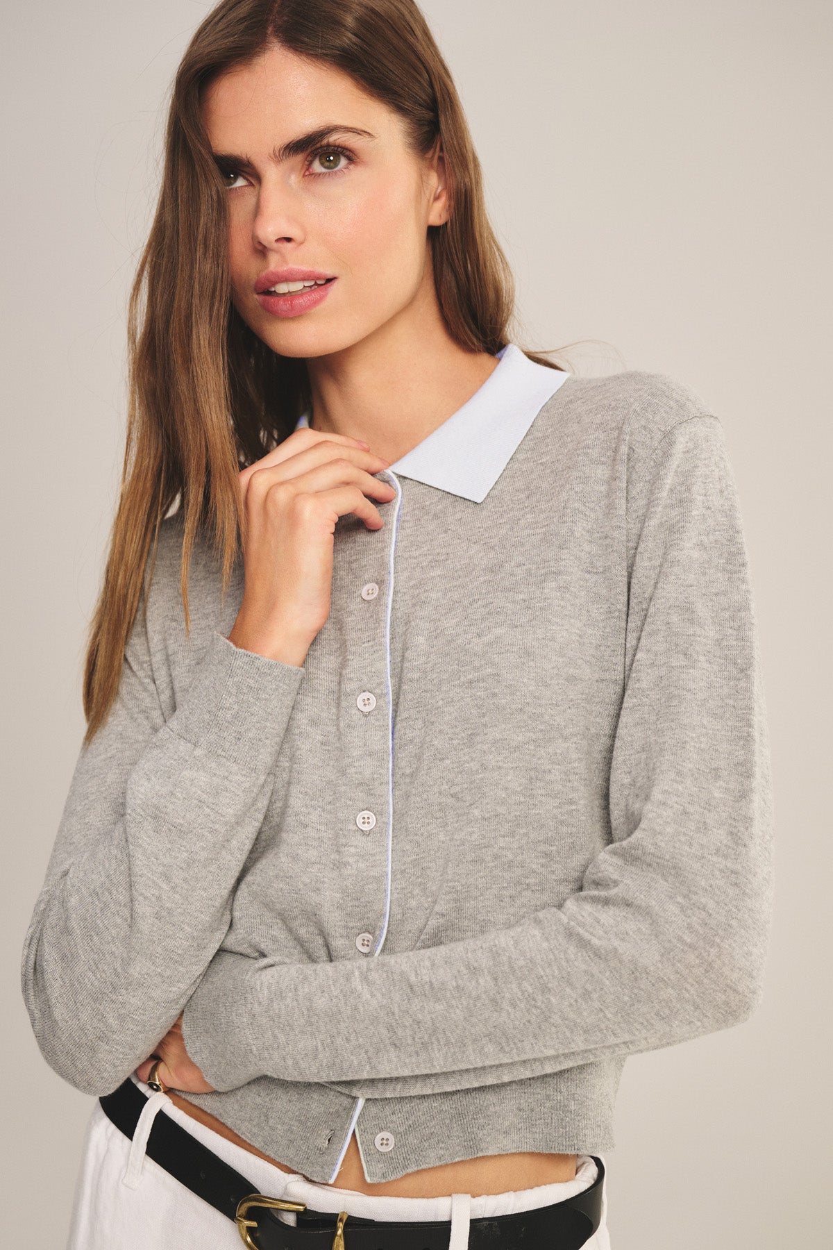 A woman stands against a plain background wearing the RIVEN CARDIGAN by Velvet by Graham & Spencer— a light gray cotton-cashmere sweater polo— layered over a light blue collared shirt, with white pants and a black belt.