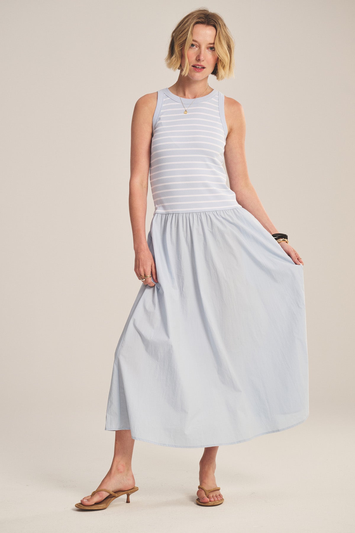 A woman wears the ROMINA DRESS by Velvet by Graham & Spencer, a sleeveless white and light gray striped cotton rib dress with a drop waist, paired with tan sandals, on a plain background.-41338485145793