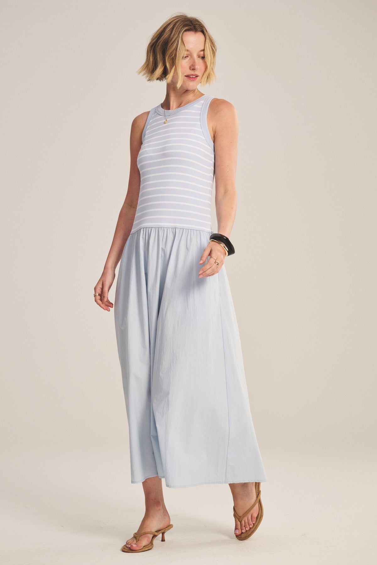 A woman wears the ROMINA DRESS by Velvet by Graham & Spencer, a sleeveless light blue and white striped cotton poplin dress, paired with heeled sandals, standing against a plain background.