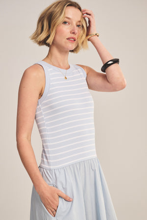 A woman with short blonde hair wears the ROMINA DRESS by Velvet by Graham & Spencer, a sleeveless light blue and white striped cotton poplin dress with a gathered skirt, accessorized with gold and black bracelets and a necklace.