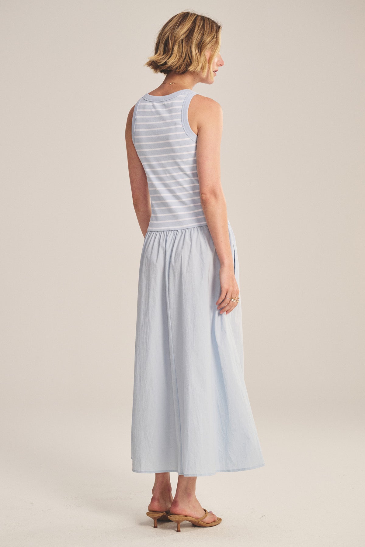 A woman faces away wearing the ROMINA DRESS by Velvet by Graham & Spencer, a sleeveless light blue and white striped midi maxi dress, paired with tan heeled sandals.